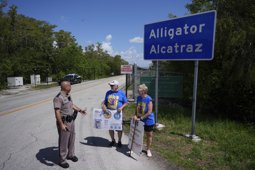 A police officer urges Art Sennholtz, 80, center, and Christy Howard, 70, of Just Us Volusia to be careful of fast-moving traffic as they hold protest signs outside the entrance to an immigration detention center in the Florida Everglades known as "Alligator Alcatraz," Thursday, Aug. 28, 2025, in Collier County, Fla. (AP Photo/Rebecca Blackwell)