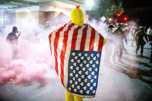 A protester, draped in an altered American flag, watches as law enforcement officers try to disperse protesters near a U.S. Immigration and Customs Enforcement facility in Portland, Ore. on Sunday, Oct. 5, 2025. (AP Photo/Ethan Swope)